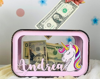 Può includere: Un salvadanaio rettangolare rosa con un fronte trasparente, contenente varie banconote da un dollaro USA. Una mano sta inserendo una banconota da un dollaro nella fessura. La scatola è decorata con un adesivo a forma di unicorno e il nome "Andrea".