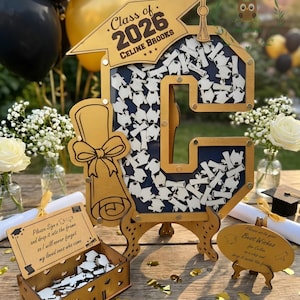 May include: A wooden graduation guest book with a large letter "G" shape, filled with small diploma-shaped notes. The design includes a graduation cap, the text "Class of 2026 Celine Brooks", and a small box for guests to sign and drop notes. Gold and navy blue accents.