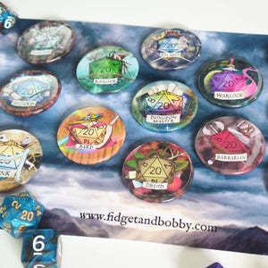 Könnte beinhalten: Eine Sammlung farbenfroher, runder, harzbeschichteter Spielmarken mit verschiedenen Dungeons & Dragons-Charakterklassen, darunter Ranger, Warlock und Bard. Jede Marke zeigt einen Würfel mit der Zahl 20 und Klassennamen. Die Website www.fidgetandbobby.com ist sichtbar.