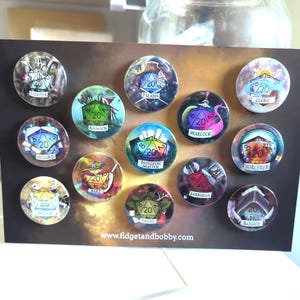 May include: A set of 12 round pins featuring different Dungeons and Dragons character classes. Each pin has a colourful illustration of a 20-sided die with a class-specific symbol and the text "20" on it. The pins are arranged in a grid on a brown background with the text "www.fidgetandbobby.com" at the bottom.
