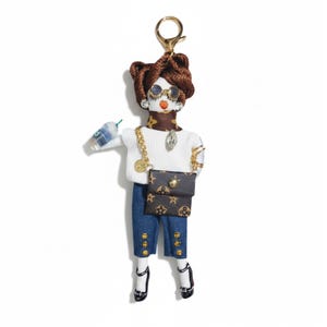 May include: A doll-shaped keychain with a brown braided hairstyle, round glasses, and a white sweater. The doll is wearing blue denim capri pants, black heels, and carrying a small purse with a brown monogram pattern. It also holds a beverage in a clear cup.