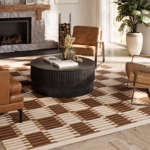 May include: A living room scene featuring a brown and beige checkered rug, two leather armchairs, and a dark round coffee table. A stone fireplace and a large potted plant add to the room's decor. The room has a warm and inviting atmosphere.