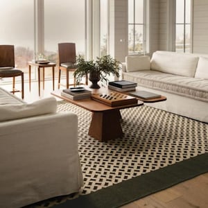 Op de afbeelding: Woonkamer met een houten salontafel, boeken en een dambord. De kamer heeft een patroonkleed, witte banken, houten stoelen en een vaas met groen. De kamer heeft grote ramen en een neutraal kleurenpalet.