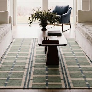 Pode incluir: Um tapete retangular com um padrão geométrico em tons de verde, branco e azul. O tapete está colocado sobre um chão de madeira, com uma mesa de centro castanha escura e dois sofás brancos. As medidas do tapete são em centímetros.
