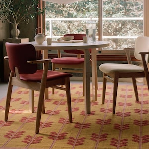 Puede incluir: Una mesa de comedor redonda de madera clara con tres sillas, dos con asientos y respaldos tapizados en rojo, y una con asiento y respaldo en color crema. Las sillas tienen marcos y reposabrazos de madera, sobre una alfombra amarilla con patrones rojos.