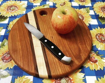 May include: A round wooden cutting board featuring a light-colored wood stripe and a circular hanging hole. A paring knife with a black handle and three silver rivets rests on the board next to a whole red and yellow apple with a green stem.