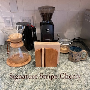 May include: A wooden napkin holder with a cherry wood finish and a natural wood stripe design. The napkin holder is labeled "Signature Stripe Cherry".