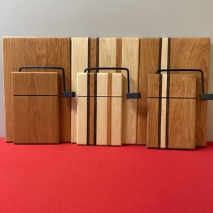 May include: Three wooden cutting boards with black metal cheese wire holders. The boards are made of different types of wood, including cherry, maple, and walnut.