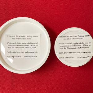 May include: Two white round containers with labels that read "Treatment for Wooden Cutting Boards and other kitchen items". The labels also include instructions for using the product, which is food grade bees wax and mineral oil. The containers are on a red surface.