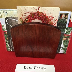May include: A dark cherry wood card holder with a curved top. The holder is filled with holiday cards. The text "Dark Cherry" is printed on a white label below the holder.