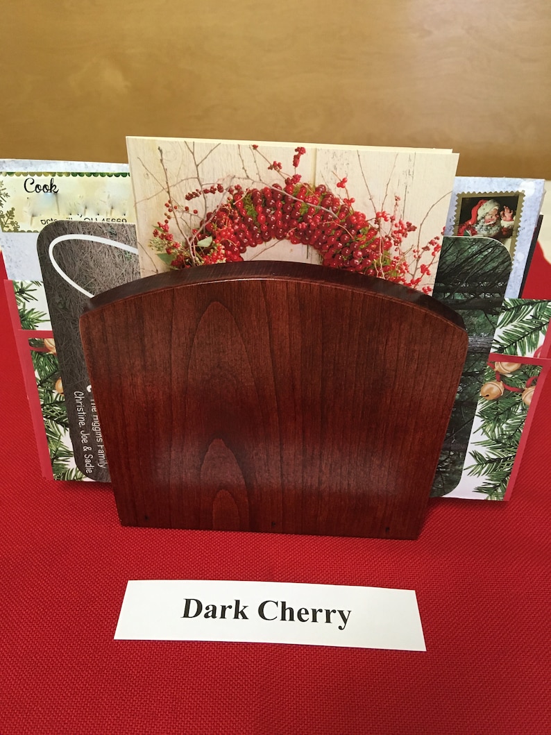 May include: A dark cherry wood card holder with a curved top. The holder is filled with holiday cards. The text "Dark Cherry" is printed on a white label below the holder.