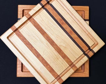May include: Two square wooden cutting boards with a raised edge and a striped pattern. The top board is larger and has a darker wood stripe in the center. The bottom board is smaller and has a lighter wood stripe in the center.