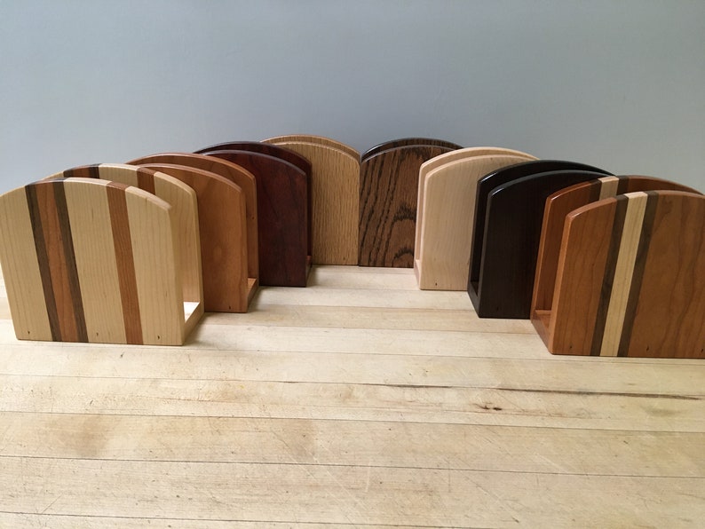 May include: A set of eight wooden napkin holders in various wood types and finishes. The holders are all the same size and shape, with a curved top and a rectangular base. The wood types include cherry, maple, walnut, and oak.