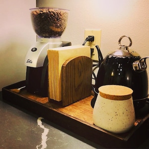 May include: A wooden tray with a black coffee grinder, a black kettle, a wooden napkin holder, and a white ceramic jar with a cork lid. The grinder has a clear container filled with coffee beans. The kettle has a silver handle and spout. The napkin holder has a wooden base and a wooden top. The jar has a white speckled design.