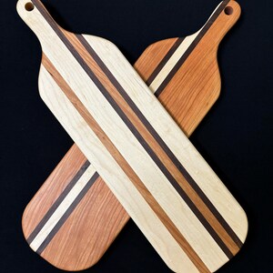 May include: Two long wooden serving boards arranged in an X shape. Each board features a striped pattern of light maple, reddish cherry, and dark walnut wood. Both boards have a rounded handle with a circular hole at the top for hanging.