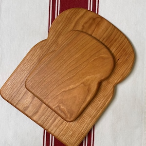 May include: Two wooden cutting boards shaped like slices of bread. The boards are made of cherry wood and have a natural finish. The smaller board is on top of the larger board.