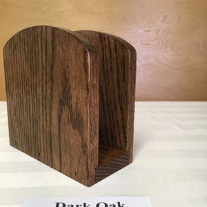 May include: A dark oak wood napkin holder with a rounded top and a rectangular base. "Dark Oak" is written on a white label below the holder.
