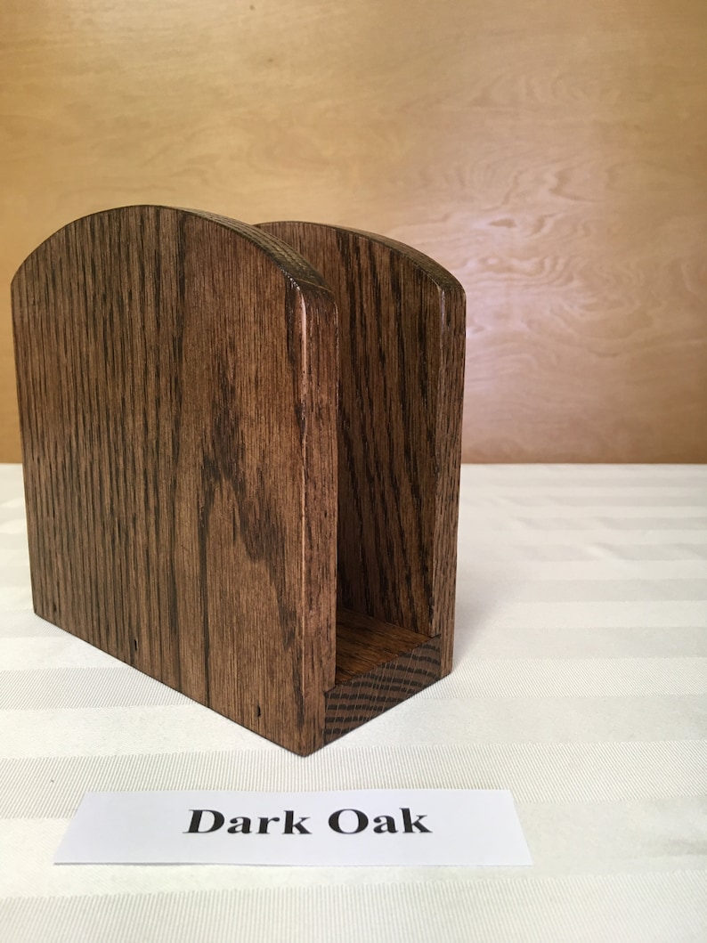 May include: A dark oak wood napkin holder with a rounded top and a rectangular base. "Dark Oak" is written on a white label below the holder.