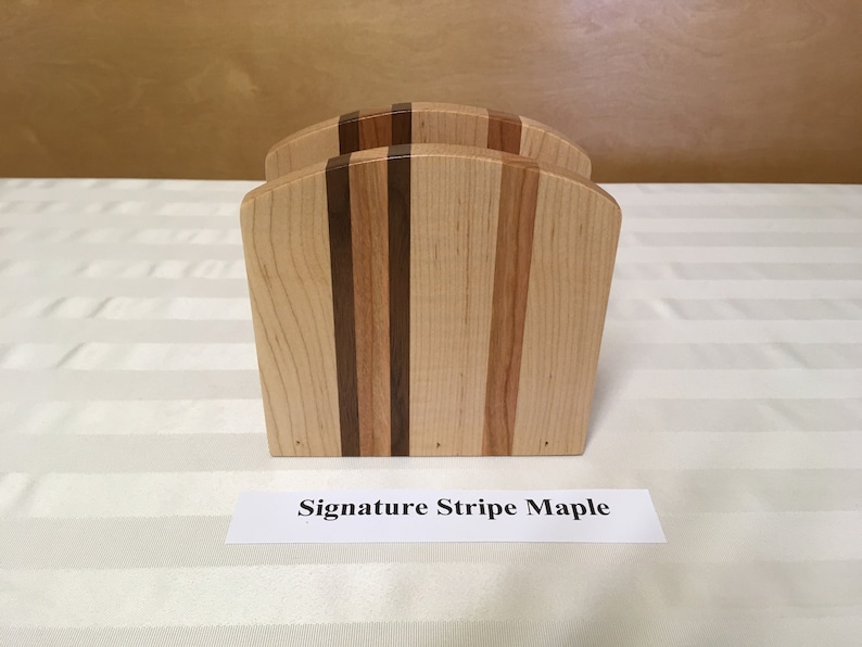May include: Wooden napkin holder with a striped design in shades of brown and maple. The holder is labeled "Signature Stripe Maple".