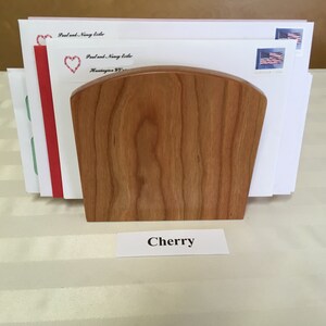 May include: A wooden letter holder made of cherry wood. The holder is holding several envelopes with stamps. The label below the holder reads "Cherry".