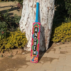 Pode incluir: Um taco de críquete colorido com uma pega azul e o nome "ANDREW" exibido verticalmente em letras cor-de-rosa. O taco apresenta um design vibrante com uma ilustração de pavão e padrões decorativos em vermelho, verde e amarelo.