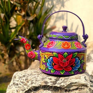 May include: A hand-painted purple teapot with a curved spout and a handle. The teapot is adorned with colorful floral designs in red, pink, orange, and blue. The teapot is sitting on a rock.