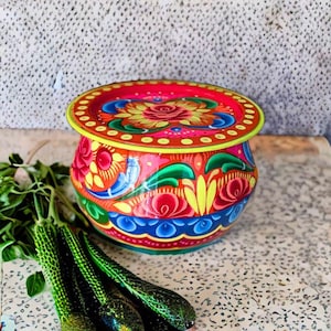 Op de afbeelding: Een fel beschilderde, ronde decoratieve container met een deksel. De container heeft een bloemenontwerp in rood, geel, blauw en groen. Het deksel is rood met gele stippen. Groene groenten staan op de voorgrond.