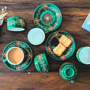 May include: A collection of green ceramic tableware with floral designs. The set includes cups, saucers, plates, and a small lidded container. The floral pattern features orange and yellow flowers with pink centers, set against a green background.