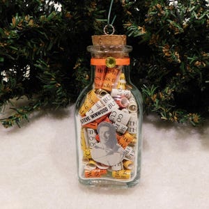 May include: A glass bottle ornament filled with rolled up pieces of paper with text, including the name "Steve Winwood". The bottle has a cork stopper and a yellow ribbon around the top. A small image of a person is visible in the bottle.