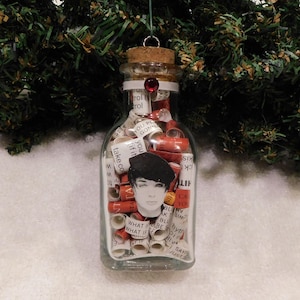 May include: A clear glass bottle Christmas ornament filled with rolled paper, red accents, and a black and white image of a person. The bottle has a cork stopper and is hanging from a green garland.