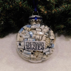 May include: Clear glass Christmas ornament filled with rolled paper and a blue label that reads "THE MOODY BLUES". The ornament has a silver cap and a blue ribbon for hanging. The background is a green Christmas tree.