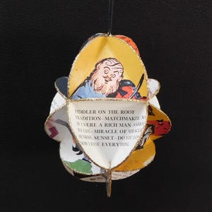 May include: A handmade, multi-faceted ornament featuring illustrations and text from "Fiddler on the Roof." The ornament has a gold glitter trim and a black string for hanging. The text includes phrases like "Tradition" and "If I were a rich man."