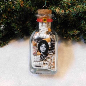May include: A clear glass bottle Christmas ornament filled with rolled paper and a black and white portrait. The bottle has a cork stopper, a red ribbon, and a gold sunflower accent. The ornament hangs from a silver loop.