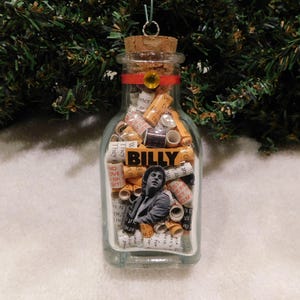 May include: A clear glass bottle ornament filled with rolled paper scrolls and a small image of a man. The bottle has a cork stopper with a red ribbon and a gold-colored gem. The word "BILLY" is visible on the bottle.