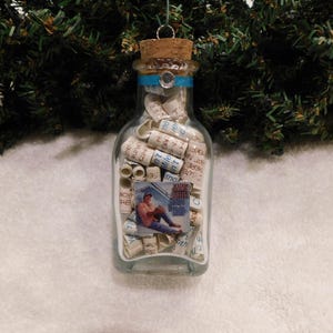 May include: A clear glass bottle ornament filled with rolled-up paper scrolls and a small picture. The bottle has a cork stopper and a blue ribbon around the neck. The ornament hangs from a silver-colored metal loop.