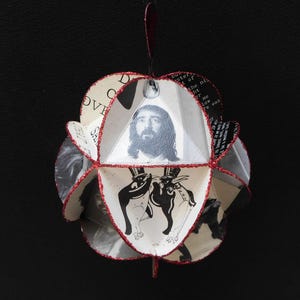 May include: A spherical ornament with a red glitter trim. The ornament features images of a man with long hair, and two figures in top hats. The background is a collage of text.