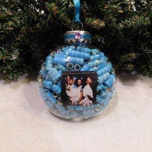 May include: Clear glass Christmas ornament filled with blue rolled paper and a photo of the Pointer Sisters. The ornament has a silver cap with a purple gem and a blue ribbon for hanging.