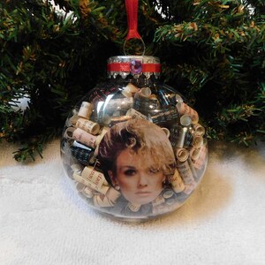 May include: Clear glass Christmas ornament filled with rolled paper and a woman's portrait. The ornament has a silver cap with a red ribbon for hanging. The background features green Christmas tree branches.