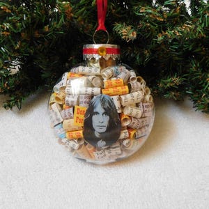 May include: Clear glass Christmas ornament filled with rolled paper and a portrait of a person. The ornament has a red ribbon for hanging and is surrounded by green Christmas tree branches.