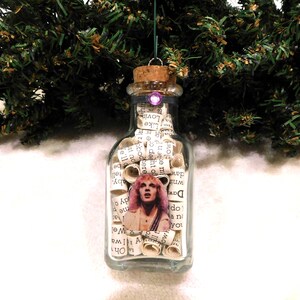 May include: A clear glass bottle ornament filled with rolled paper and a picture of a person with pink hair. The bottle has a cork stopper and a purple gem on top. The ornament is hanging in front of a green Christmas tree.