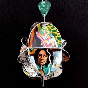 May include: A unique pendant necklace featuring a layered design with images of a guitar, a person, and other graphics. The pendant is made from recycled materials and has a green guitar pick at the top for hanging.