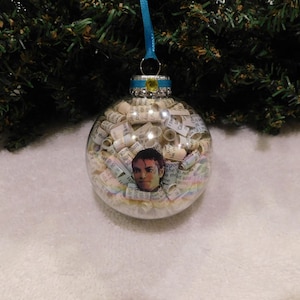 May include: Clear glass Christmas ornament filled with rolled paper and a portrait of a person. The ornament has a blue ribbon for hanging and is decorated with small gems.