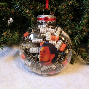 May include: A clear glass Christmas ornament filled with rolled up pieces of paper with words printed on them. The ornament has a red ribbon and a silver top. A black and white photo of a man is visible through the paper.