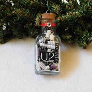 May include: A clear glass bottle ornament filled with rolled paper and a black and white U2 logo. The bottle has a cork stopper with a red ribbon and a metal eyelet for hanging. The ornament is suspended in front of a green Christmas tree.