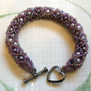 May include: A purple and silver beaded bracelet with a heart-shaped clasp. The bracelet is made with small seed beads and larger pearl-like beads.