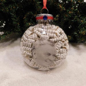 May include: A clear glass Christmas ornament filled with rolled up pieces of paper with text. The ornament has a red and blue ribbon and a blue jewel at the top. A man's face is visible through the paper.