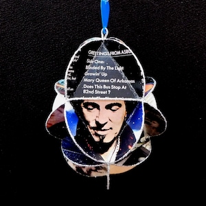 May include: A handmade ornament featuring a portrait of a man, with a blue ribbon for hanging. The ornament is made from recycled materials and includes song titles like "Blinded By The Light".