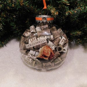 May include: Clear glass Christmas ornament filled with rolled paper and a photo of a person with long blonde hair. The ornament has the words "LED ZEPPELIN" on it and is hanging from a green Christmas tree.