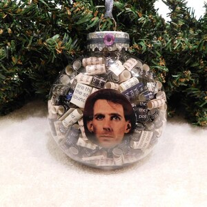 May include: Clear glass Christmas ornament filled with rolled paper and a portrait. The ornament has a silver cap with a purple gem and a silver ribbon for hanging. The ornament is surrounded by green pine needles.
