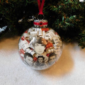 May include: A clear glass Christmas ornament filled with rolled up paper scraps with text, and small pictures of people. The ornament has a red ribbon and a red bow at the top.